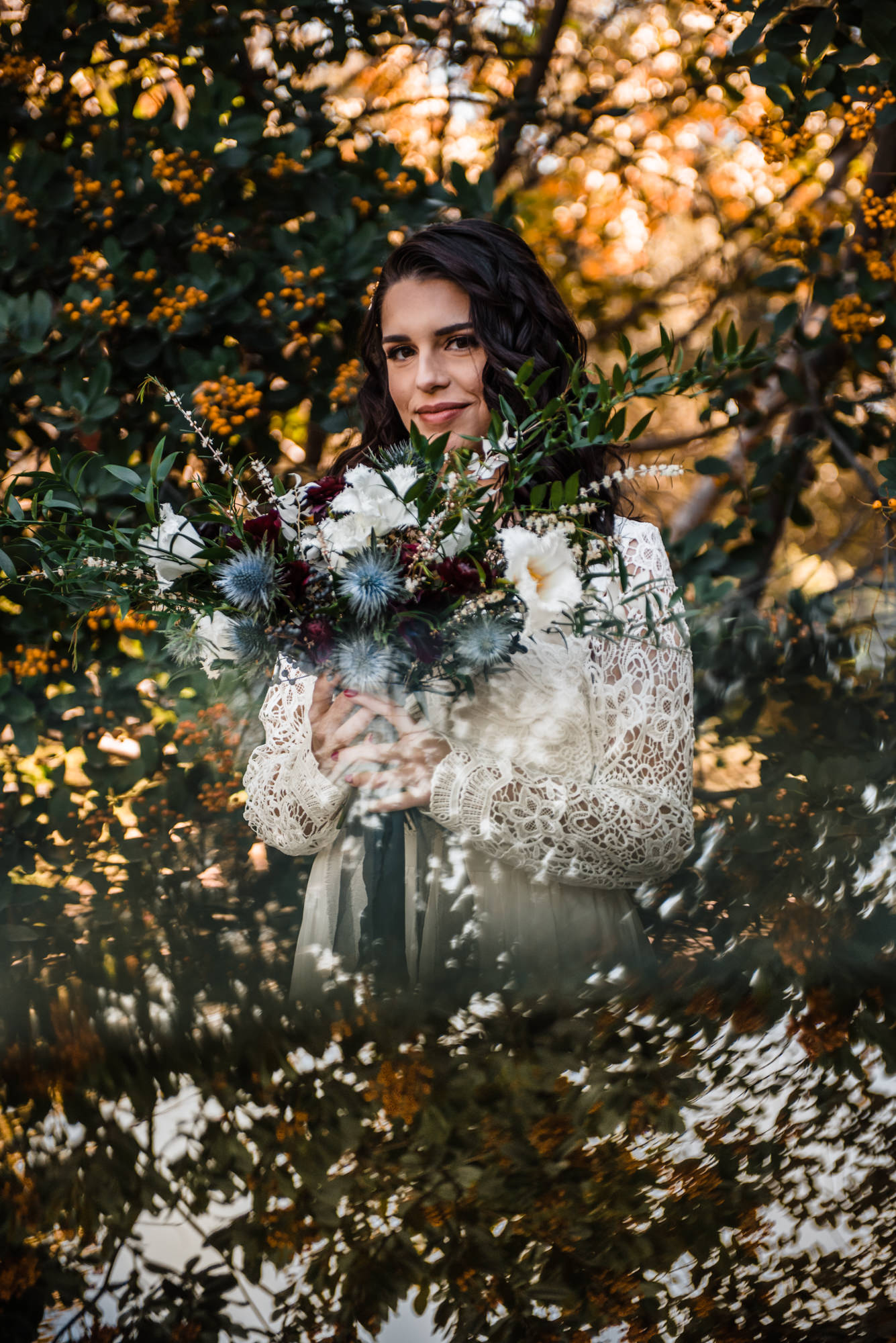 bride stands surrounded by trees in her lace wedding dress holding a large bouquet of blue and white flowers 