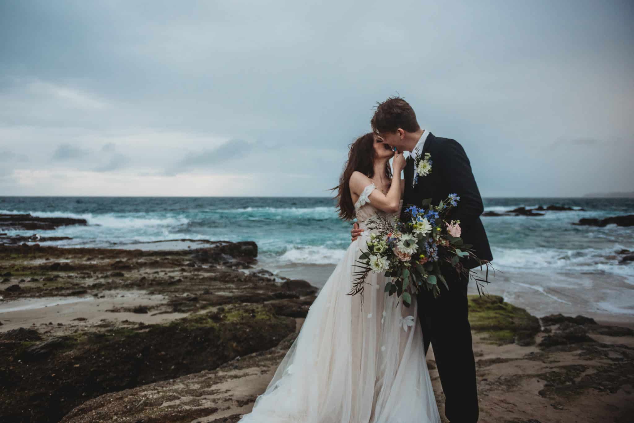 bride in off-shoulder wedding dress kisses her new husband while her holds the bouquet in front of the crashing waves by the sea