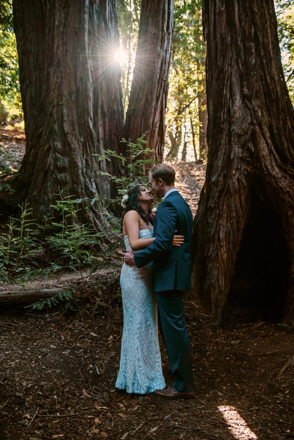 newly eloped couple kisses among tall trees while the sun peeks through following their elopement