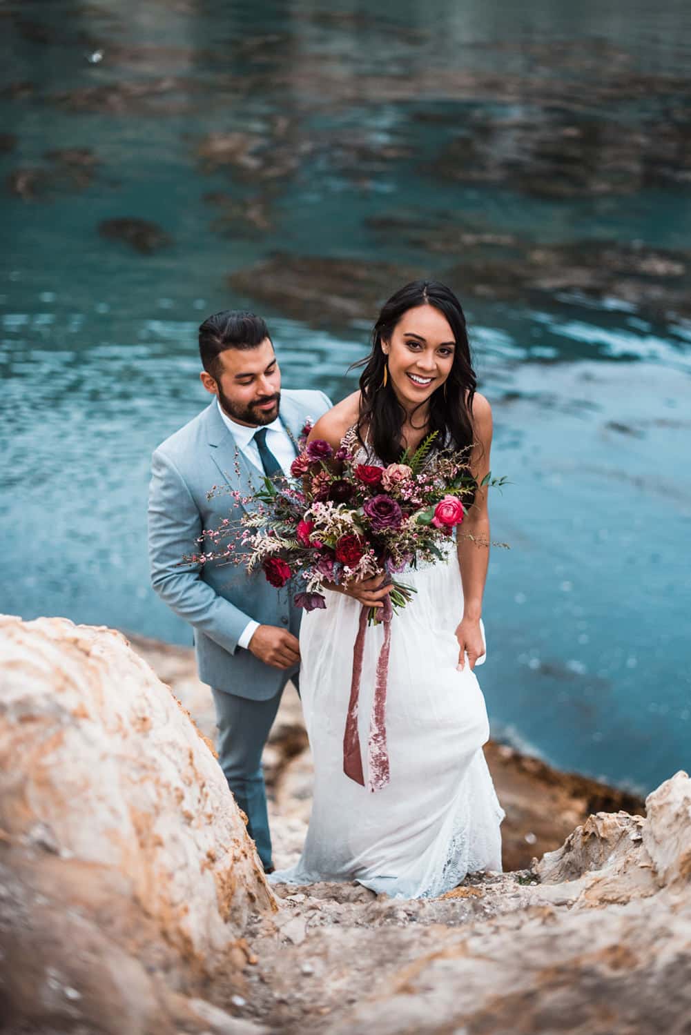 groom stands behind bride as she walks up rocky pathway holding a large bouquet of burgundy flowers
