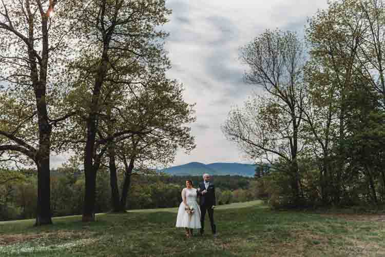 A bride and groom standing on a green hill in new england with forest in the background