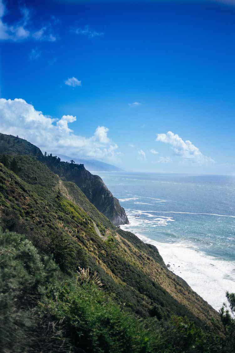 cliffside view of Big Sur with the green rocky hillside on one half and the blue Pacific ocean on the other on a clear day