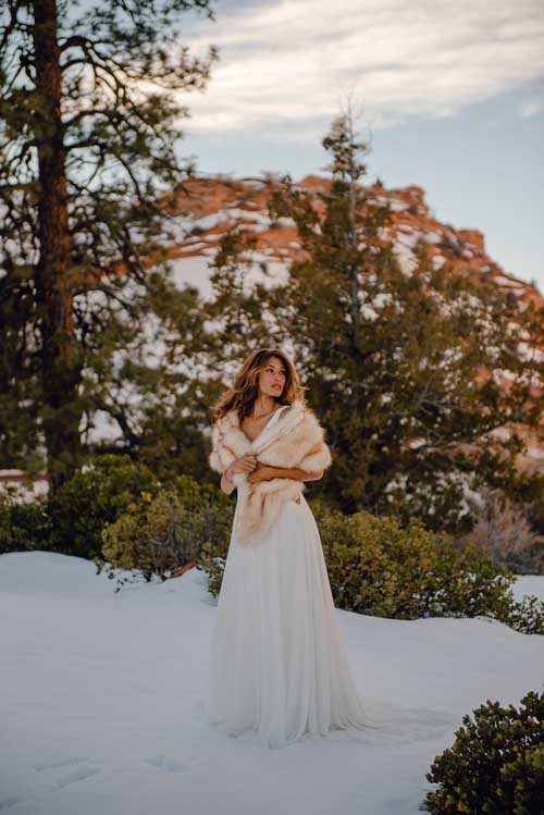 bride stands in the snow in her wedding dress with faux fur wrapped around her shoulders to keep her warm