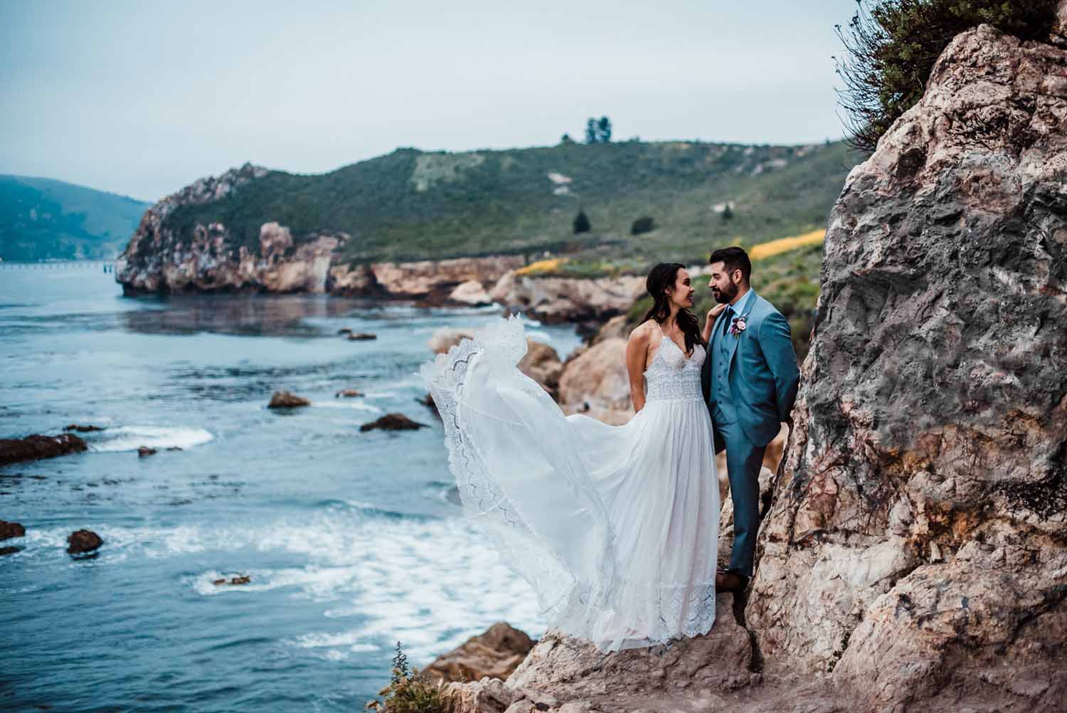 bride wearing the perfect elopement dress of sheer spaghetti strap and lace blowing in the wind while she and her groom stand oceanside perched on a cliff