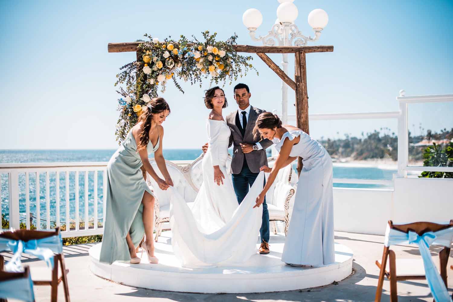 two bridesmaids fix the bride's dress while she stands at the seaside outdoor altar with her husband