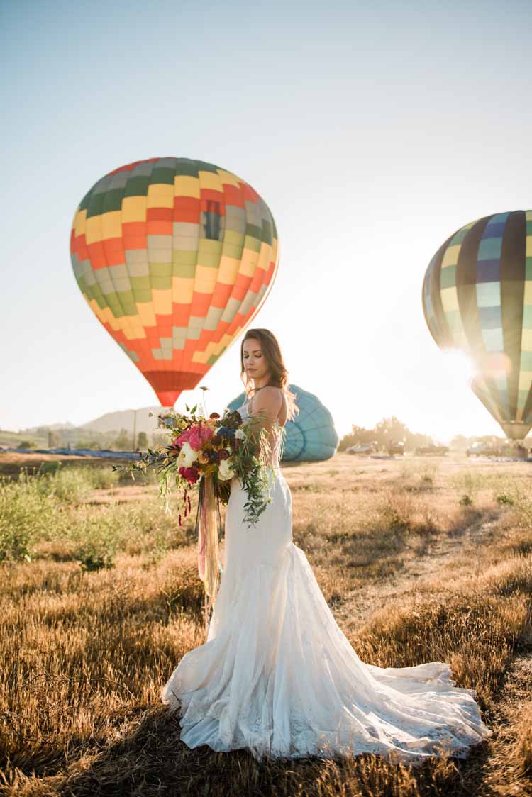 bride stands in a field in her spaghetti strap wedding gown at sunset with hot air balloons rising in the background