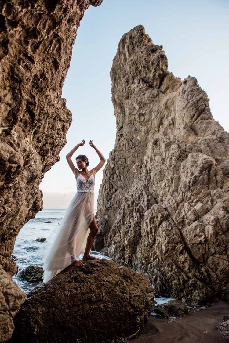 bride standing on beach framed by two large rocks with her hands up dancing in her sheer wedding dress