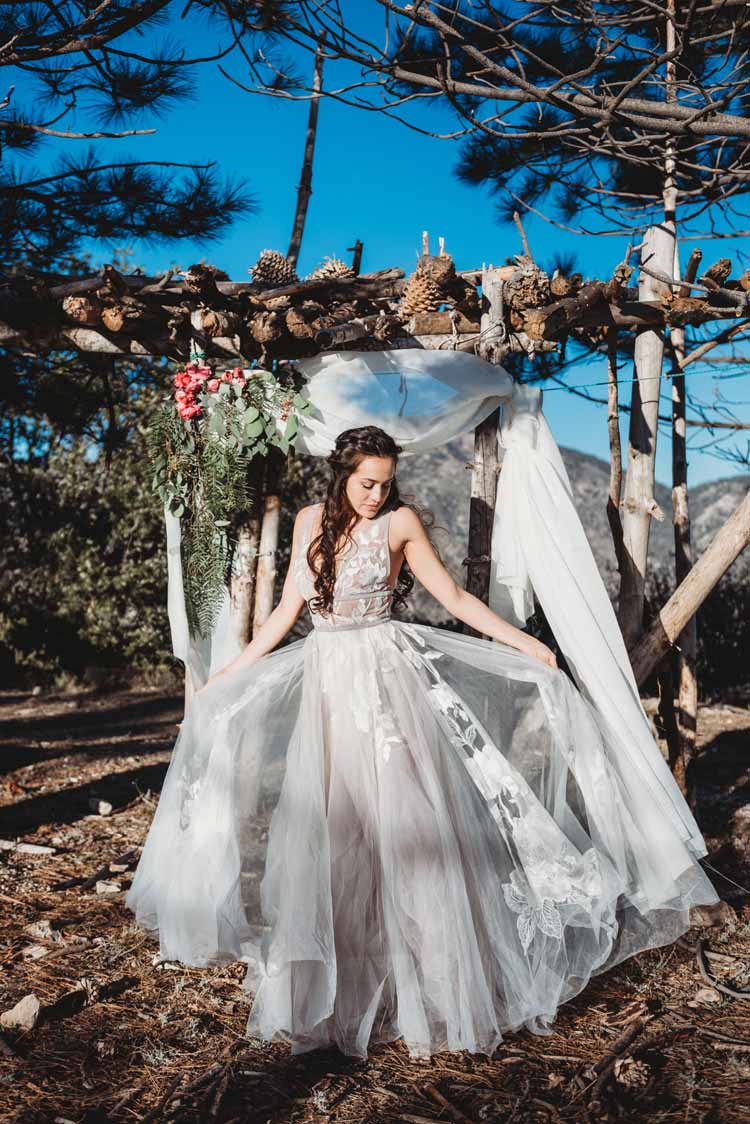 bride stands under a tree while holding up either side of her sheer wedding dress to show off the details