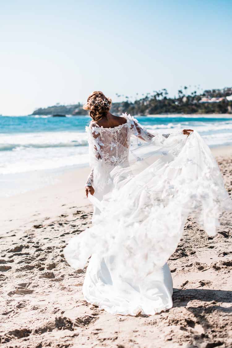 bride strolls down the beach facing away from the camera while her lace wedding elopement dress flows in the wind