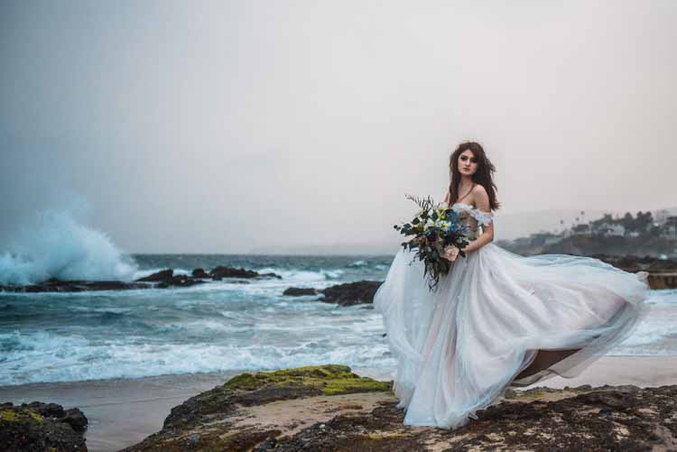 bride stands on the beach with her off-shoulder wedding dress flowing in the wind