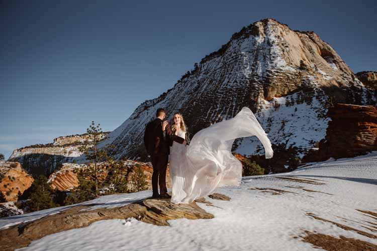 bride's wedding gown flows in the wind atop a hill with a mountain view while standing with her groom
