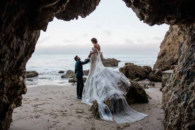 bride in flowing white dress stands on top of a rock while looking down at her husband on their wedding day in front of the ocean