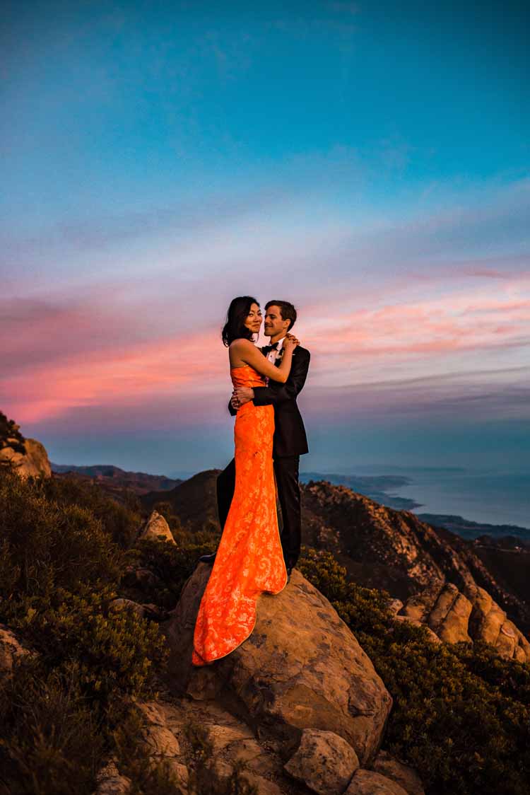 bride smiles in her red wedding dress while embracing her new husband at sunset on a rocky beach