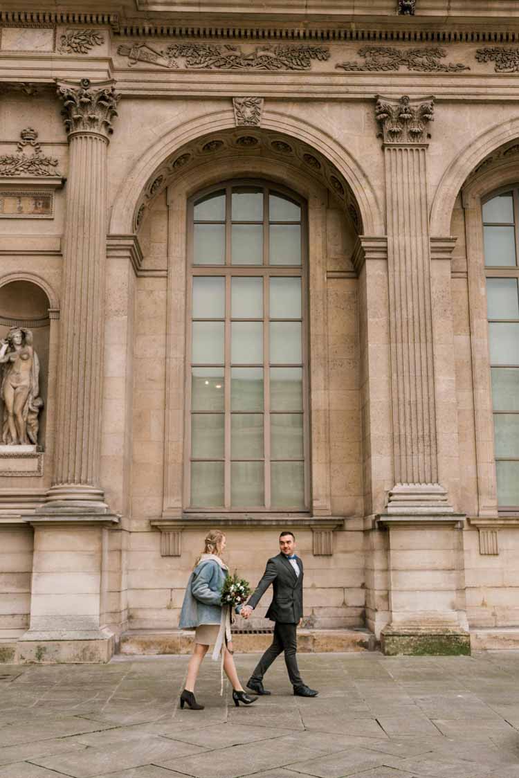 newly engaged couple strolls holding hands in front of an old building for their Paris engagement during the winter