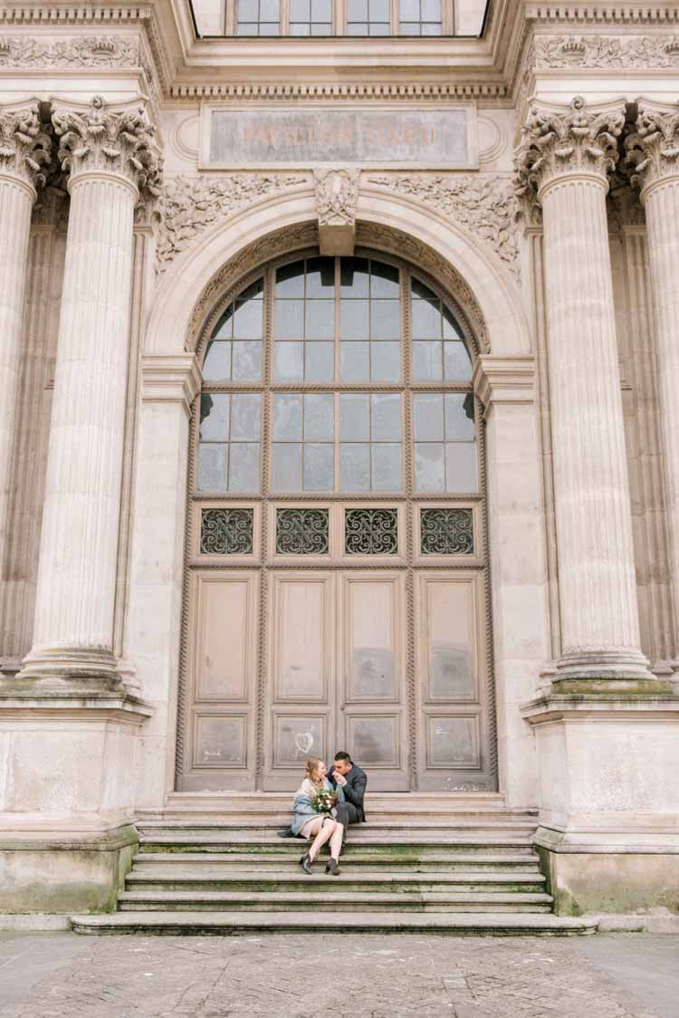 couple sits on steps in front of an old building during their Paris engagement shoot while the man kisses his fiance's hand