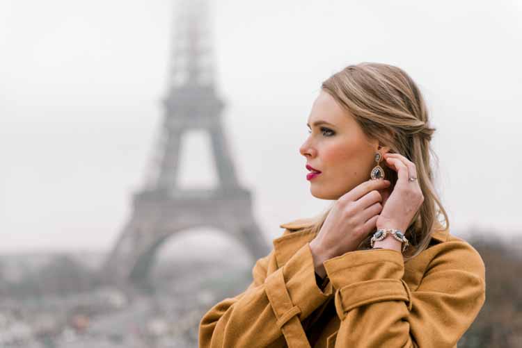 newly engaged woman clasps earrings closed while looking out into Paris during the winter