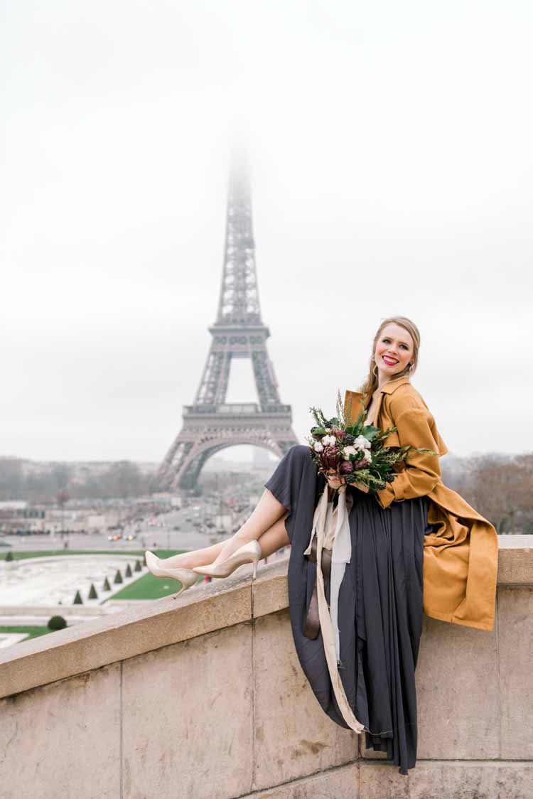 woman sits on a ledge in her grey dress holding a bouquet during her Paris engagement photoshoot 