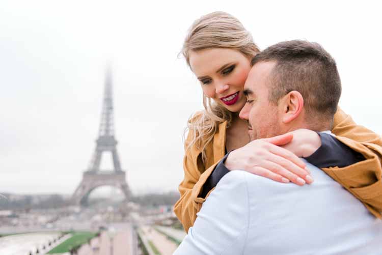 future married couple embraces in Paris in front of the Eiffel Tower to celebrate their engagement while smiling