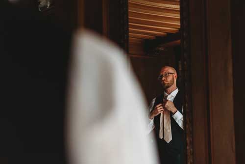 A groom preparing his tie while looking in the mirror before an elopement