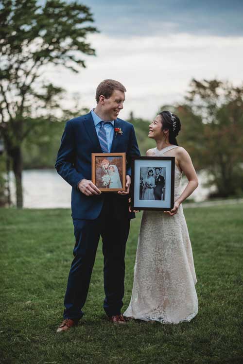 A bride and groom holding portraits while standing on a green lawn with a lake in the background during an elopement