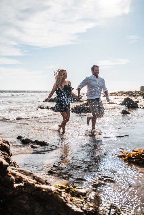 A bride and groom running and smiling on the beach shore during an elopement