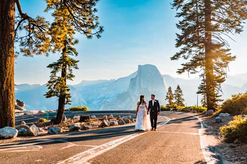 A bride and groom holding hands while walking on a road with yosemite in the background