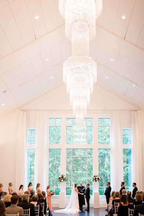 A bride and groom being married in a white venue with large windows in the background and large chandelier over top during an elopement