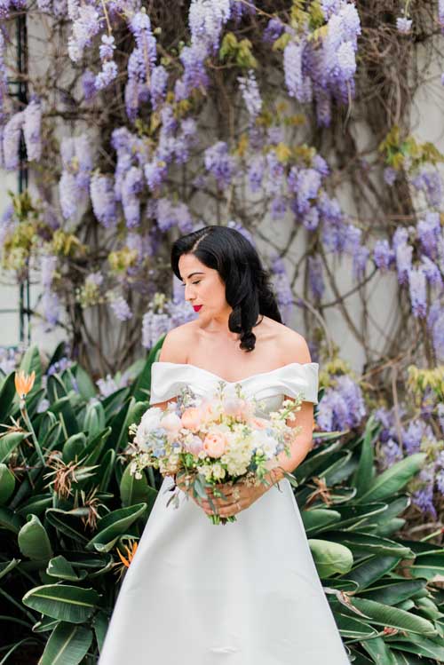 A bride in a white dress holding a bouquet with green and purple plants in the background during an elopement