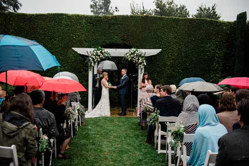 A bride and groom being married with a green grass wall in the background with family present holding umbrellas during an elopement