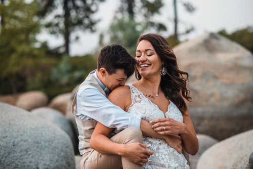 A groom hugging and kissing a bride from behind while sitting on large rocks during an elopement