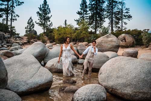 A couple laughing while stepping between rocks on the watery shore of lake tahoe during an elopement